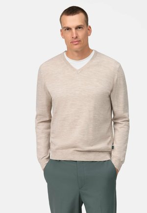 Beige V-neck sweater made of textured knit fabric, paired with a white T-shirt underneath, worn with dark green trousers.