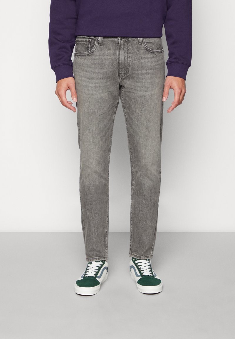 Jeans de mezclilla gris con un corte recto, cinco bolsillos y textura desgastada. Combinados con zapatillas verdes y blancas. Sudadera morada visible.