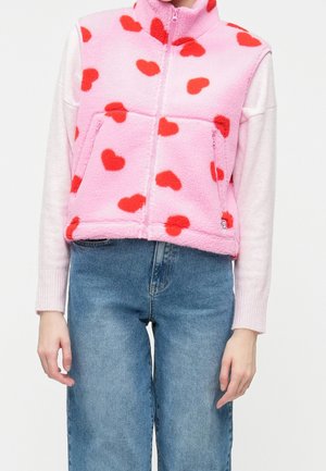 Persoon draagt een roze fleecevest met rode hartpatronen over een lichtroze shirt met lange mouwen en blauwe spijkerbroek, staand tegen een witte achtergrond.