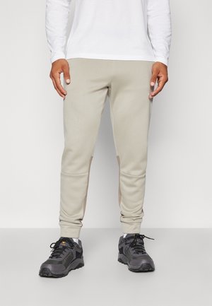 Man draagt beige slim-fit joggers met geribde boorden, gecombineerd met zwarte en grijze sportschoenen en een wit shirt met lange mouwen, staand tegen een witte achtergrond.