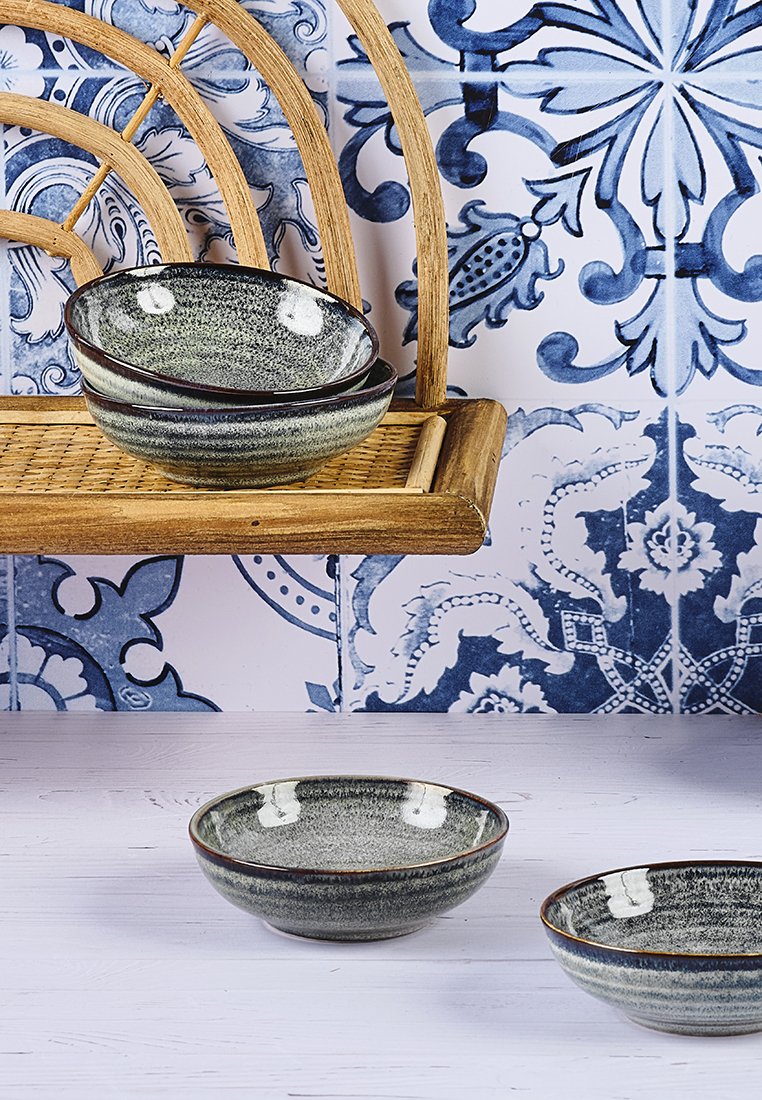Cuencos de cerámica con un acabado moteado, color gris oscuro y bordes marrones. Están apilados en una estantería de madera frente a una pared de color azul con estampado.