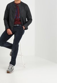 Uomo seduto su uno sgabello bianco, indossando una giacca di pelle nera, una camicia borgogna, pantaloni scuri e sneaker grigie, su uno sfondo bianco semplice.