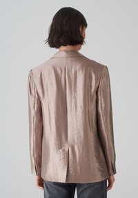 Metallisch roségoldener Blazer mit glatter Textur, Reverskragen, geschlitztem Rücken und langen Ärmeln, kombiniert mit dunklen Jeans.