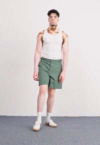 Camiseta blanca sin mangas, pantalones cortos verdes y zapatillas blancas con suela de goma. Fondo sencillo, individuo en posición neutral.