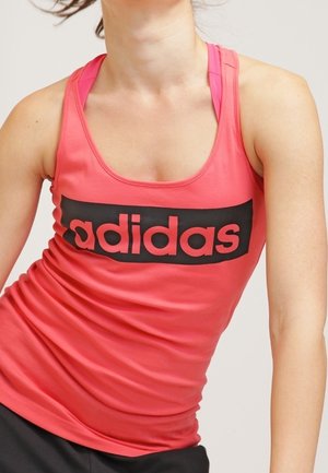 Nainen pukeutunut istuvaan punaisen väriseen Adidasin hihattomaan toppiin, jossa musta logo, näyttäen ylävartalon ja osan mustista housuista yksinkertaisella taustalla.