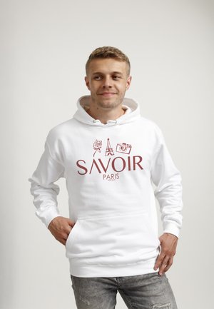 Ung mand iført en hvid hoodie med teksten "SAVOIR PARIS", rose, Eiffeltårnet og kameraikoner, står med hænderne i lommerne.