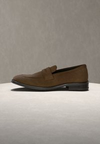 Mocassin homme en daim marron avec semelle noire et petit talon empilé sur surface claire et fond neutre.