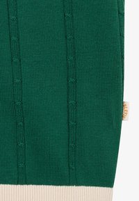 Tela de punto verde con patrones de cables verticales y un dobladillo acanalado color crema, que presenta una pequeña etiqueta crema con texto naranja en el costado.