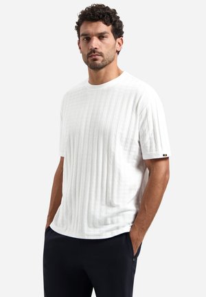 Man met donker krullend haar, gekleed in een wit getailleerd shirt met korte mouwen en een zwarte broek, staand met handen in de zakken tegen een effen achtergrond.