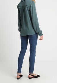 Blusa in teal a maniche lunghe con cucitura raccolta sul retro, abbinata a jeans a pantalone skinny blu e ballerine nere con taglio sul retro.