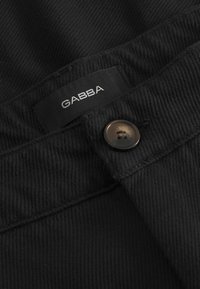 Gros plan sur un tissu en denim noir avec une étiquette GABBA, un bouton marron et une couture texturée le long de la taille.