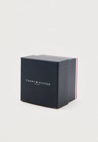 Tmavě modrá kartonová krabička s matným povrchem. Na přední straně se nachází bílý logotyp "TOMMY HILFIGER" a text "WATCHES", s akcentovými barvami na bočních okrajích.