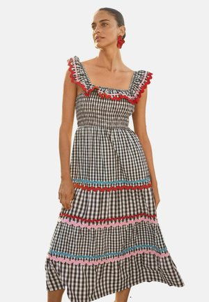 Kvinde iført en sort-hvid gingham-kjole med rynkede kanter i rødt, pink og blåt, stående foran en ensfarvet baggrund.