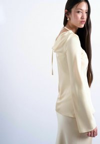 Jeune femme aux longs cheveux foncés portant un chemisier en soie crème à nouer dans le dos et une jupe assortie, debout devant un fond blanc uni.