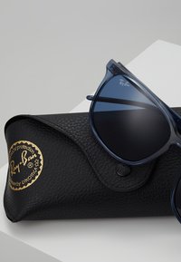Ochelari de soare Ray-Ban din plastic bleumarin, cu lentile întunecate, expuși lângă un toc negru din piele texturată cu brandul aurit.