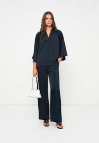 Navyseide Oberteil mit weiten Ärmeln und V-Ausschnitt, kombiniert mit passenden weiten Hosen. Das Modell trägt eine kleine weiße Handtasche. Schwarze Sandalen runden den Look ab.