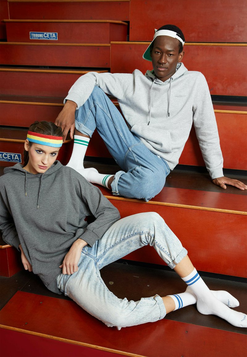 Dwa modele siedzą na drewnianych schodach. Jeden z nich ma na sobie szary hoodie, jasnoniebieskie dżinsy i wielokolorową opaskę na głowę. Drugi ma na sobie szary hoodie, jasne dżinsy oraz białe skarpetki w zielone paski.