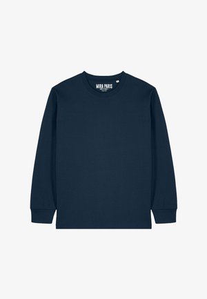 Žena s rovnými čiernymi vlasmi, oblečená v tmavomodrej dlhorukávovej košeli, voľných modrých džínsoch a bielych teniskách, stojaca pred jednoduchým bielym pozadím.