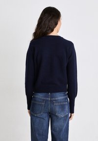 Navy gestrickter Pullover mit gerippten Details, Rundhalsausschnitt und leicht verkürztem Schnitt, kombiniert mit dunkelblauen Jeans aus Denim mit aufgesetzten Taschen.