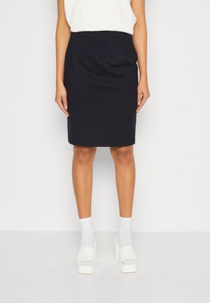 Femme portant une jupe crayon noire longueur genou, un haut blanc, des chaussettes blanches et des sandales à plateforme blanches, debout devant un fond blanc uni.