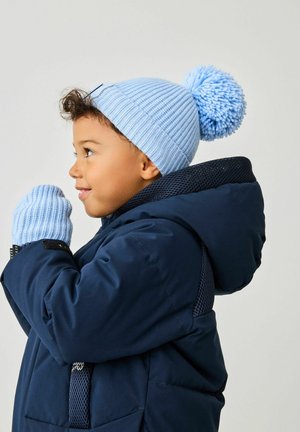 Jasnoniebieska dzianinowa czapka z pomponem, o ribbed texture, w zestawie z pasującymi jasnoniebieskimi rękawiczkami. Noszona na granatowej pikowanej kurtce.