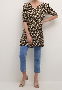Luipaardprint tuniek met korte pofmouwen, knoopdetail en een ontspannen pasvorm, gecombineerd met lichtblauwe cropped jeans en beige muiltjes.