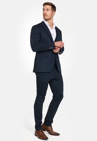 Marineblauer Anzug mit passender Hose, maßgeschneiderte Passform und strukturierter Stoff, kombiniert mit einem weißen Hemd und braunen Dressschuhen.