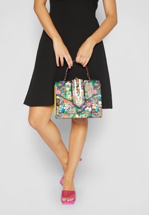 Kobieta w czarnej sukience trzymająca kolorową florystyczną torebkę z złotym łańcuszkiem, nosząca różowe buty na obcasie z odkrytymi palcami, stojąca na jednolitym tle.