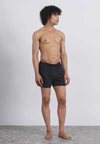 Jauns vīrietis ar lokainiem matiem stāv basām kājām, ģērbies melnās šortos pret vienkāršu gaiši pelēku fonu, skatoties pa kreisi.