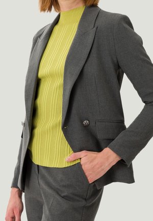 Osoba ubrana w szary, dopasowany garnitur z dwurzędową marynarką i pasującymi spodniami, w połączeniu z prążkowanym, limonkowo-zielonym golfem.