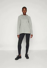 Sivi dukserica s kapuljačom i vezicama, u paru s crnim tajicama s teksturiranim detaljima i crnim sportskim cipelama.