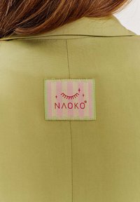 Tkanina w oliwkowym odcieniu z tkanym wzorem ma wyraźną naszywkę marki NAOKO w różowym i zielonym, prezentującą proste grafiki i tekst.