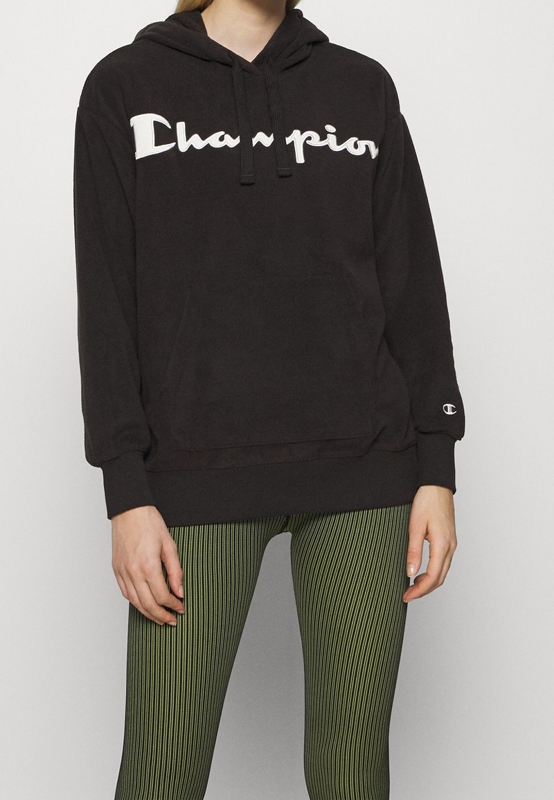 Personne portant un sweat à capuche noir Champion avec un logo blanc et des leggings rayés verts, se tenant devant un fond clair uniform.