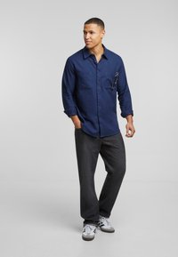 Camisa de manga larga azul marino con dos bolsillos en el pecho, combinada con vaqueros oscuros y zapatillas grises, con un diseño sencillo y casual.