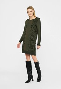 Robe à manches longues vert olive avec un motif léopard, en tissu tricoté, coupe droite, associée à des bottes noires à talons montant jusqu'aux genoux.