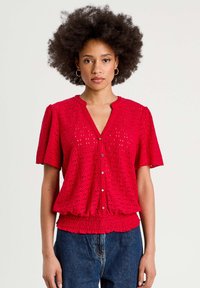 Blouse rouge à manches courtes avec un motif texturé, dotée d'une fermeture à boutons et d'une taille élastique pour une coupe décontractée.