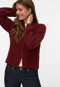 Burgundijska zip-up kardigan sa rebrastim rukavima i teksturiranim tijelom, uparena s tamnim trapericama. Ima opušteni kroj i suptilni sjaj.