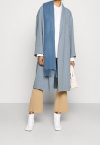 Persona che indossa un cappotto texturizzato azzurro chiaro, una sciarpa blu, una camicia bianca, pantaloni beige a gamba larga, stivaletti bianchi e tiene in mano una borsa bianca.