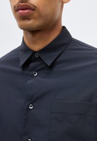 Κοντινό πλάνο του λαιμού και του στήθους ενός άνδρα που φοράει σκούρο μπλε πουκάμισο με κουμπιά, μυτερό γιακά και τσέπη στο στήθος.