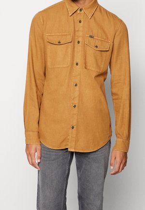 Mann trägt ein senfgelbes langärmeliges Hemd mit zwei Brusttaschen und graue Jeans vor einfachem Hintergrund.