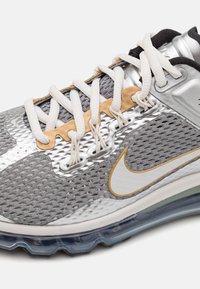 Grå och silver atletisk sko med vita snörningar, guldaccenter, överdel i mesh och genomskinlig luftkuddesula på vit bakgrund.