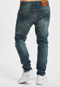 Modré džíny z denimu s úzkym strihom, s jemným vyblednutím, detailom na poškodených kolenách a klasickými zadnými vreckami. Hnedá kožená nášivka na páse.