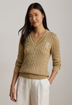 Femme portant un pull beige en maille torsadée à col en V avec un monogramme sur la poitrine et un pantalon blanc taille haute, debout avec une main dans la poche.