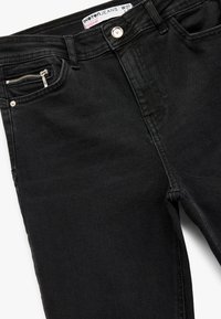 Jeans de mezclilla negra con un corte ajustado. Presenta un bolsillo frontal con un zipper plateado, cierre de botón metálico y una etiqueta en la cintura.