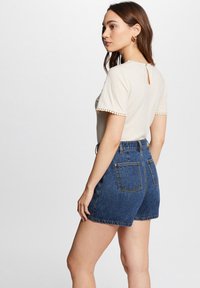 Top crema con bordes de mangas acampanadas y escote en la espalda, combinado con shorts de mezclilla azul de tiro alto que cuentan con bolsillos traseros y detalles dorados.