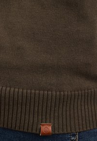 Brązowy sweter dzianinowy z ryflowanym wykończeniem, z małą brązową skórzaną metką w prawym dolnym rogu. Teksturowana tkanina i prosty design.