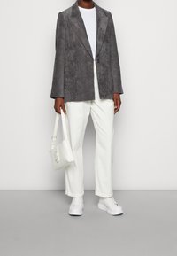 Blazer in velluto a coste grigio con rever a punta, abbinato a una t-shirt bianca, pantaloni bianchi e sneaker bianche. Tiene in mano una piccola borsa bianca.