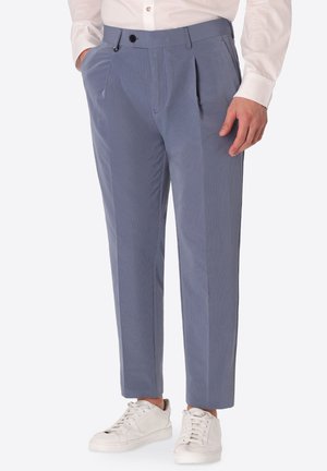 Uomo che indossa pantaloni blu plissettati con tasche laterali, abbinati a sneakers bianchi e una camicia bianca a maniche lunghe con bottoni.