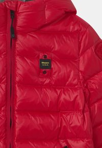 Rode pufferjack met een geborduurd ontwerp, voorzien van een capuchon en een zwart etiket met gele tekst. Gladde, glanzende stof en ritssluiting.
