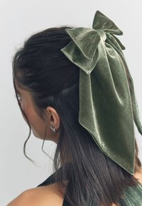 Green velvet hair bow with long, flowing tails attached to dark, straight hair. Bow features a textured surface and outlined edges.